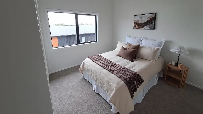 Neutral-toned bedroom with a wide black aluminium window overlooking neighbouring residential buildings.