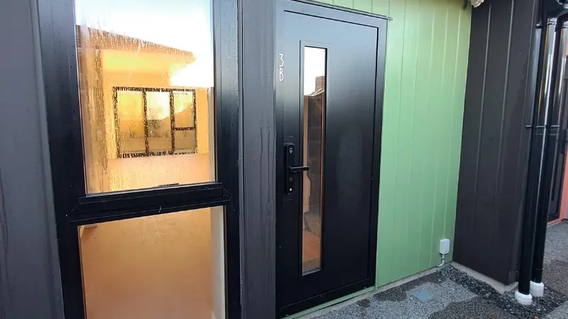 Dark aluminium entrance door with narrow vertical glazing beside a large stairwell window on a green-clad townhouse.