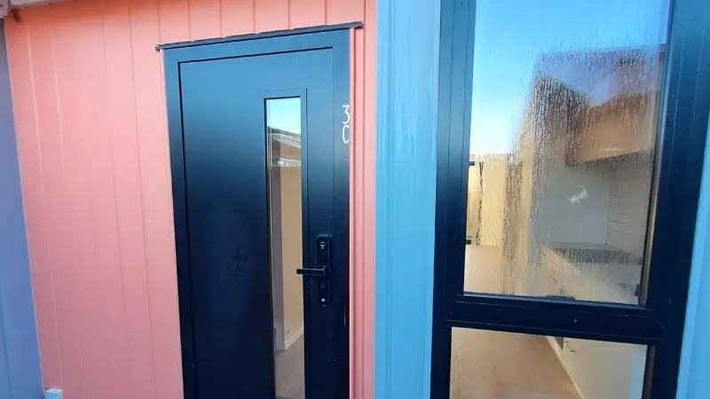 Black aluminium entrance door with narrow glazing beside a large window on a coral and blue townhouse exterior.