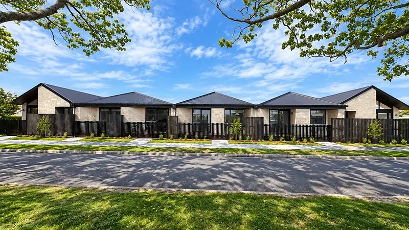 Row of single-storey brick townhouses at 299 Levers Road, Matua, Tauranga, behind dark slatted fencing under mature trees