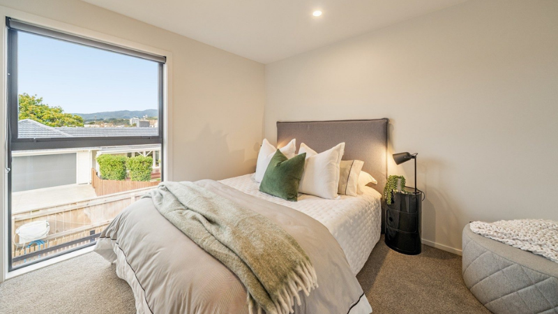 Styled bedroom with a large dark-framed aluminium window overlooking neighbouring homes and distant hills.
