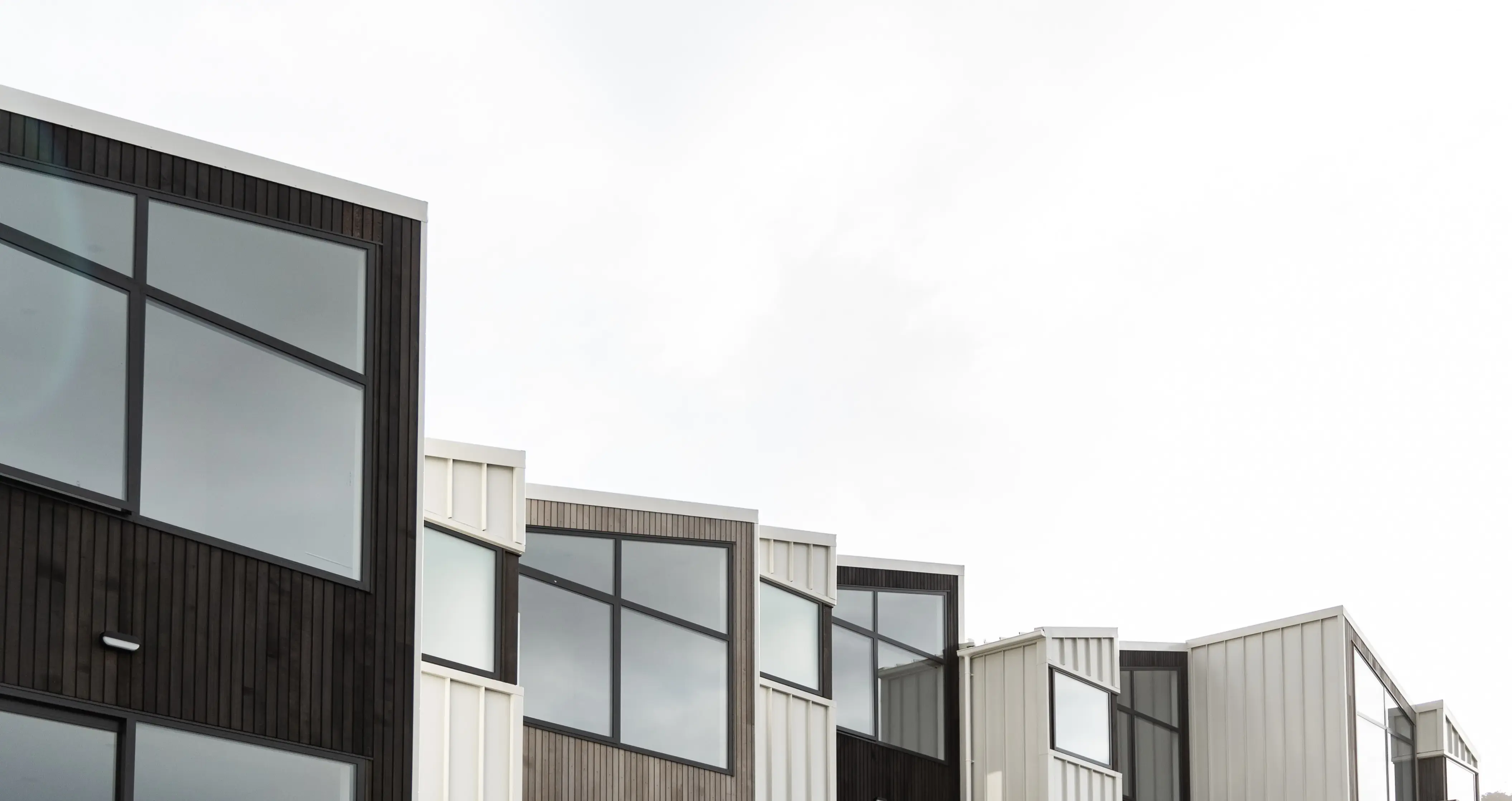 A row of modern multi-residential townhouses featuring large aluminium windows, vertical cladding in black, grey, and white tones, and staggered rooflines under a bright overcast sky.