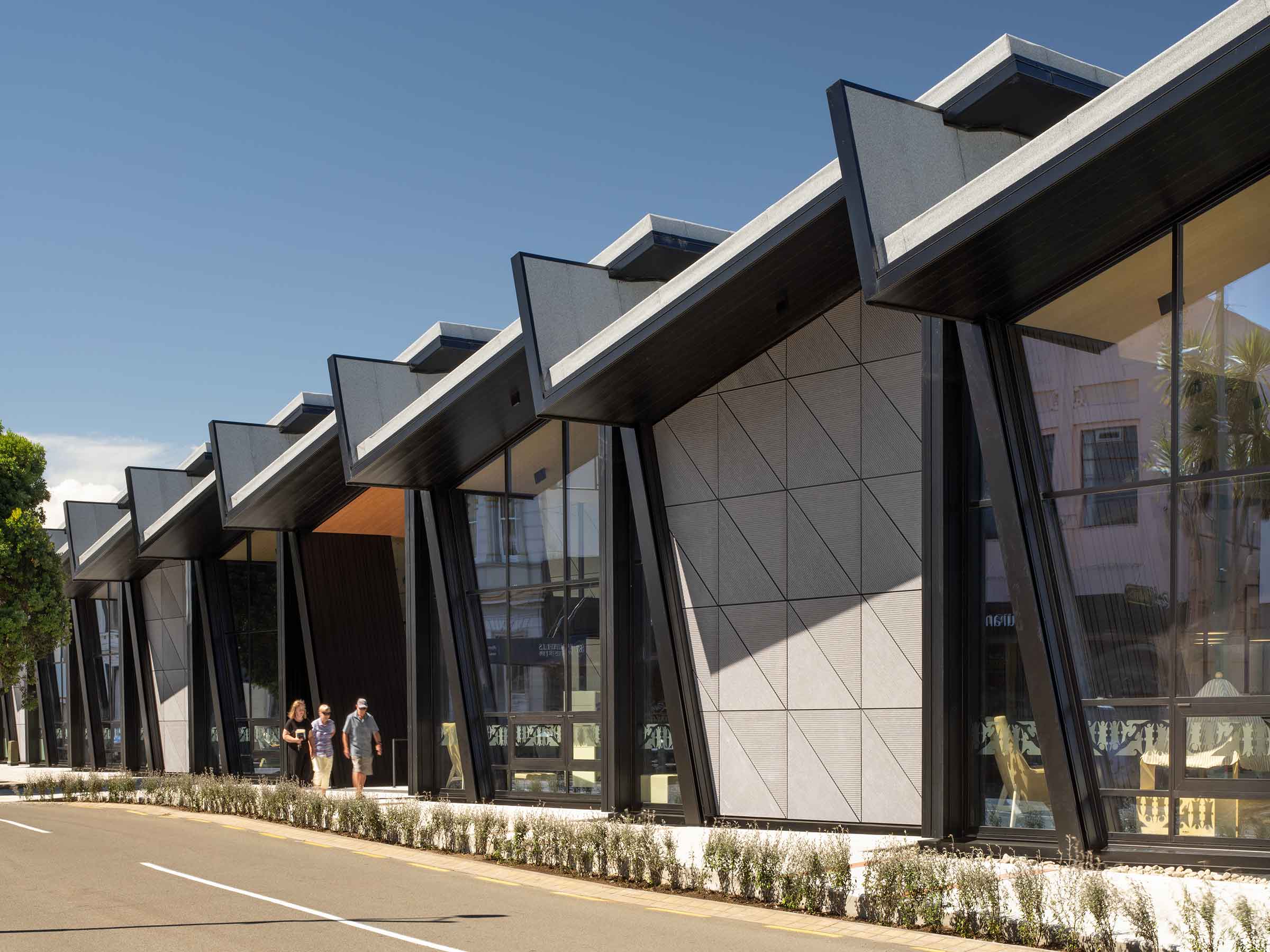 Side view of Te Ramanui o Ruapūtahanga’s exterior featuring angular sawtooth roofing, flushglaze panels, and dark aluminium framing with pedestrians walking by.