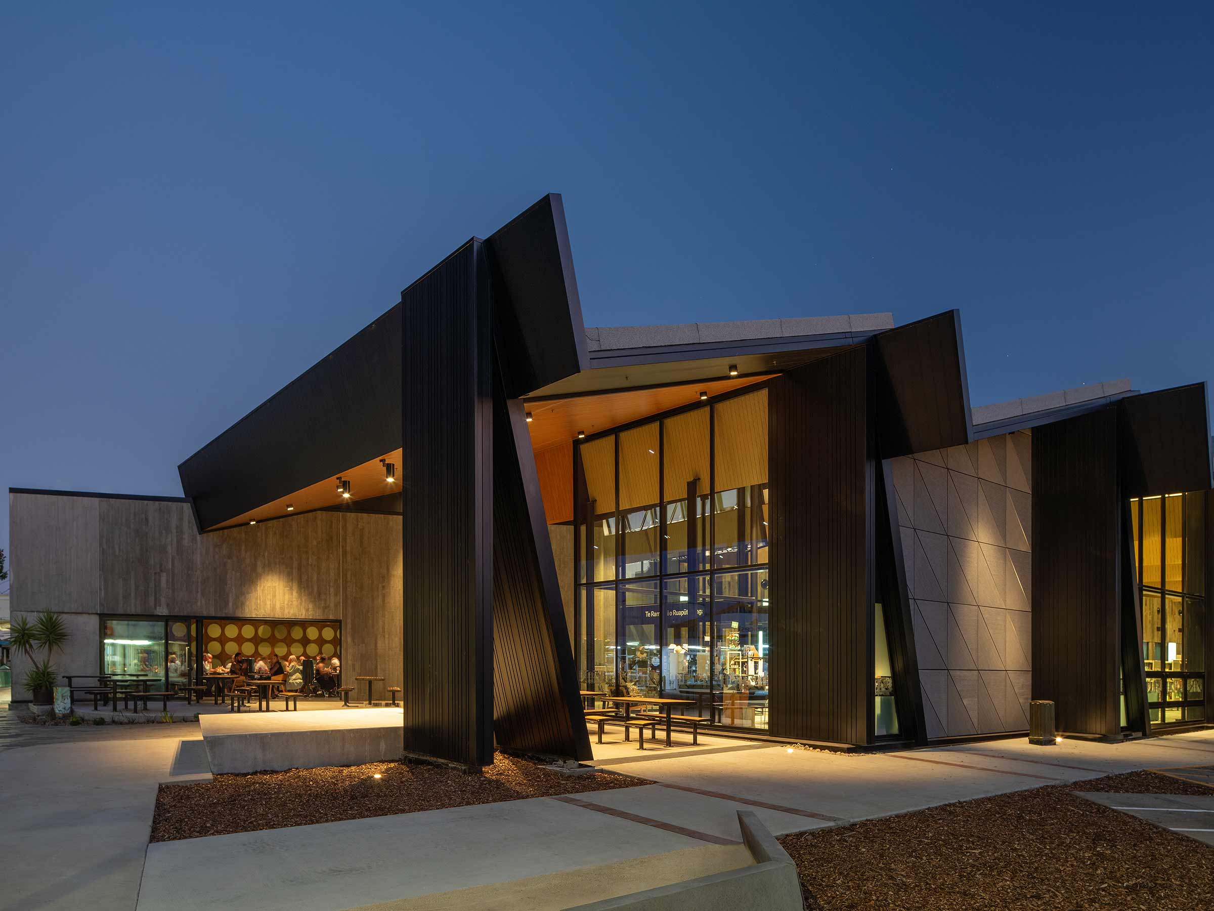 Contemporary architectural building at dusk featuring bold angular rooflines, large glass facades, and a mix of textured materials including dark metal cladding and concrete panels. Warm interior lighting glows through the expansive windows, while outdoor seating areas and minimalist landscaping accentuate the modern design.