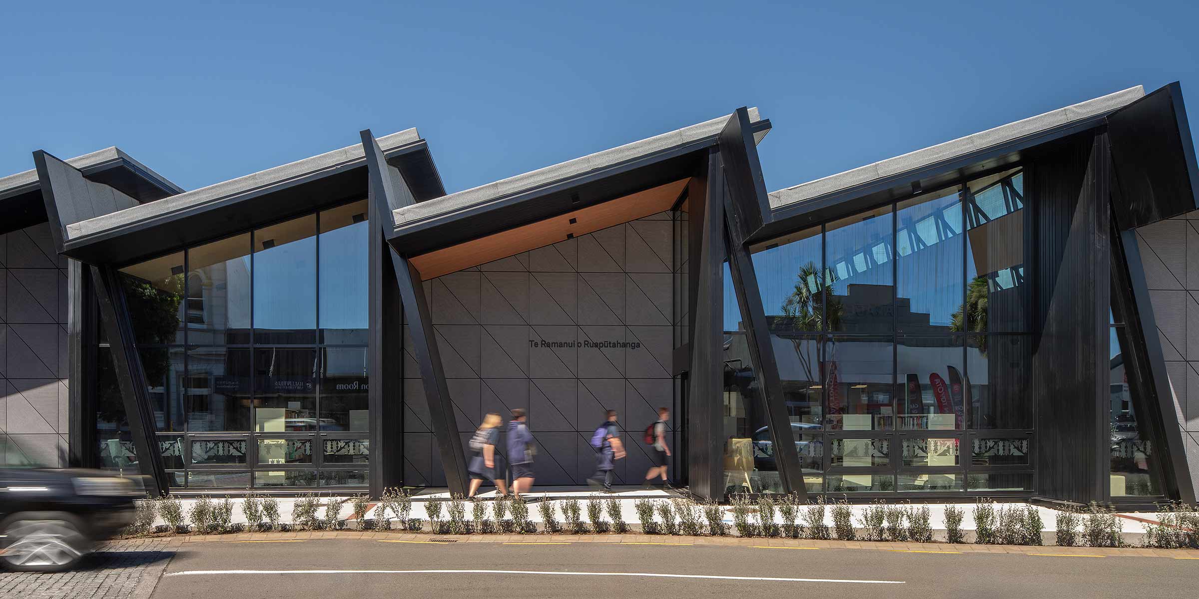 Modern public building with a striking zigzag roofline and large floor-to-ceiling windows framed in dark metal. The façade features geometric concrete panels, while pedestrians walk along the street in front, and a blurred car passes by, conveying urban movement under a clear blue sky.