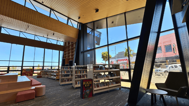 Interior view of the Te Ramanui o Ruapūtahanga Community Centre showing tall glazed façades with aluminium joinery, bookshelves, and timber ceiling panels flooding the space with natural light.
